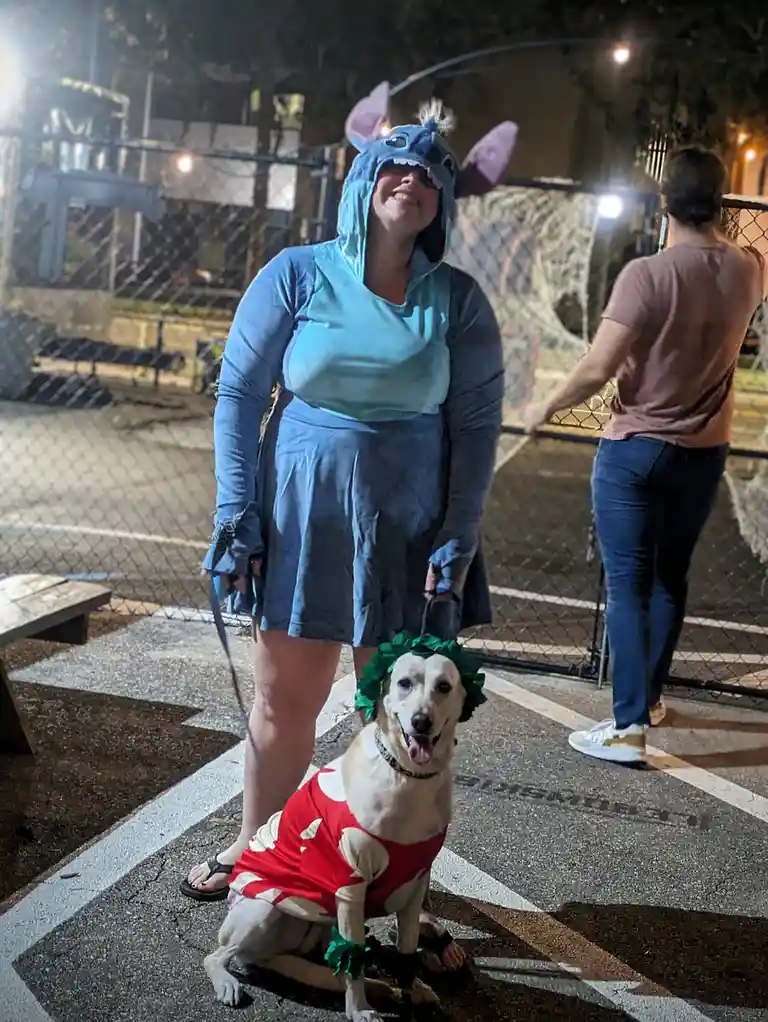 A person in a blue Stitch costume smiles while holding a leash attached to a dog wearing a red and white outfit with a green…