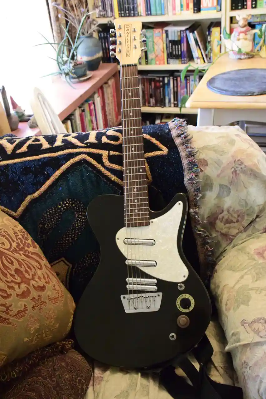 A black Danelectro electric guitar with a white pickguard rests upright on a floral-patterned couch, surrounded by patterned…