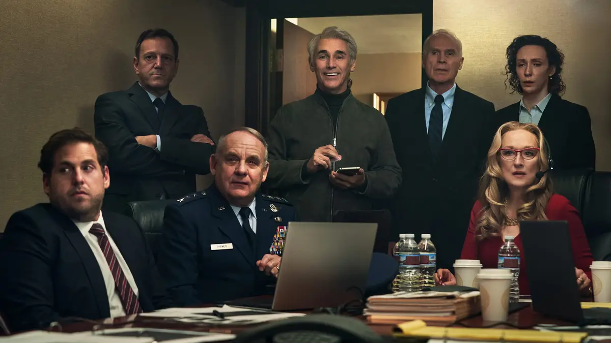 A group of formally dressed people are gathered in a tense-looking conference room, with one man in a green jacket standing…