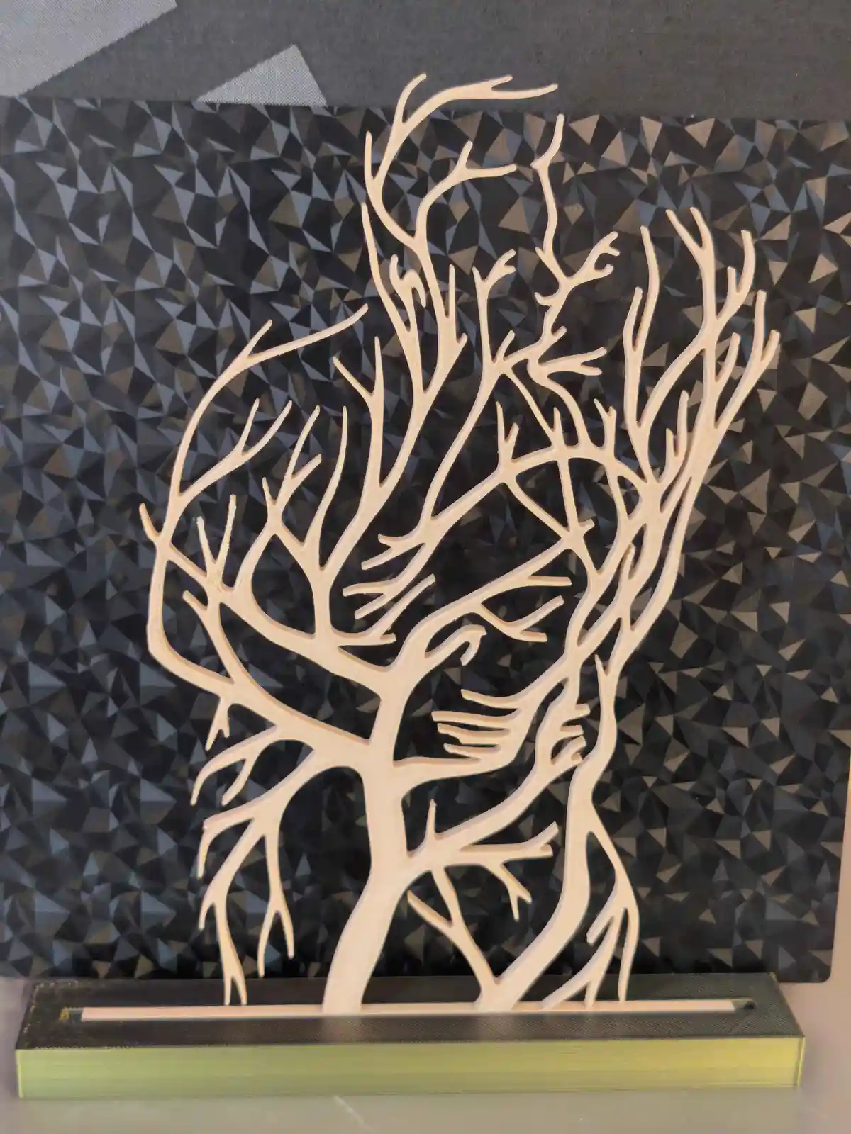A light-colored, stylized tree sculpture with intricate, branching limbs is mounted on a dark, textured geometric…