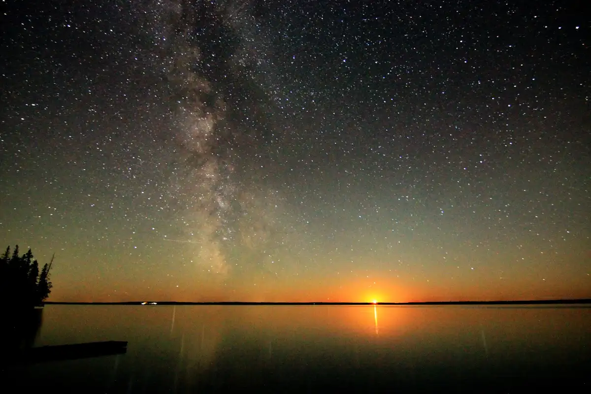 A stunning night sky photograph shows the Milky Way galaxy arching across a star-filled sky above a calm body of water. On…