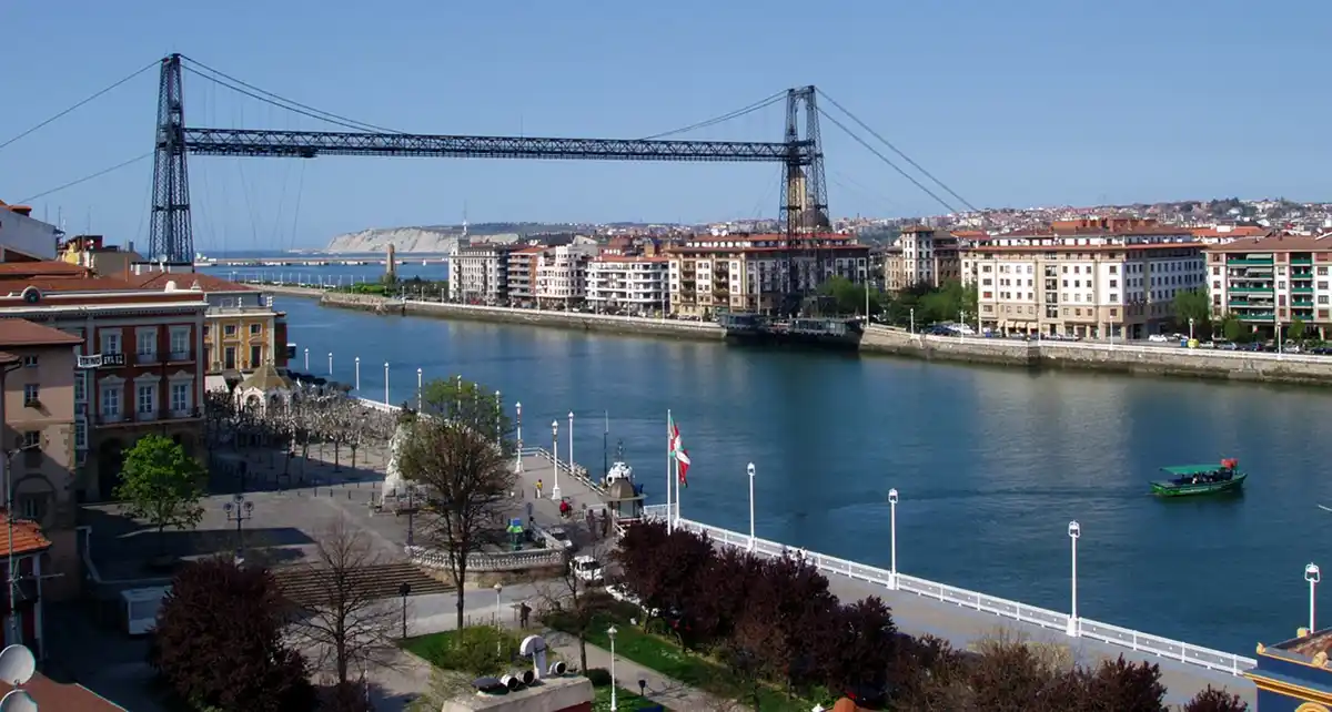 A scenic view of a city with a large suspension bridge spanning a river, flanked by multi-story buildings with red-tiled…