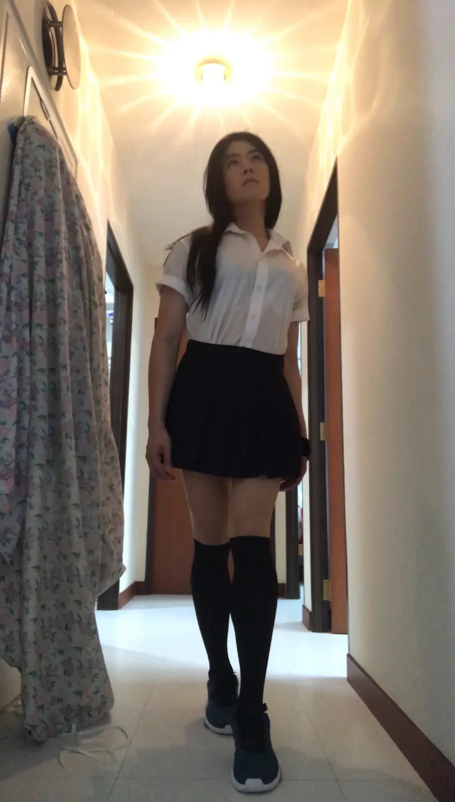 A person stands in a hallway wearing a white short-sleeved shirt, a black pleated skirt, black knee-high socks, and…