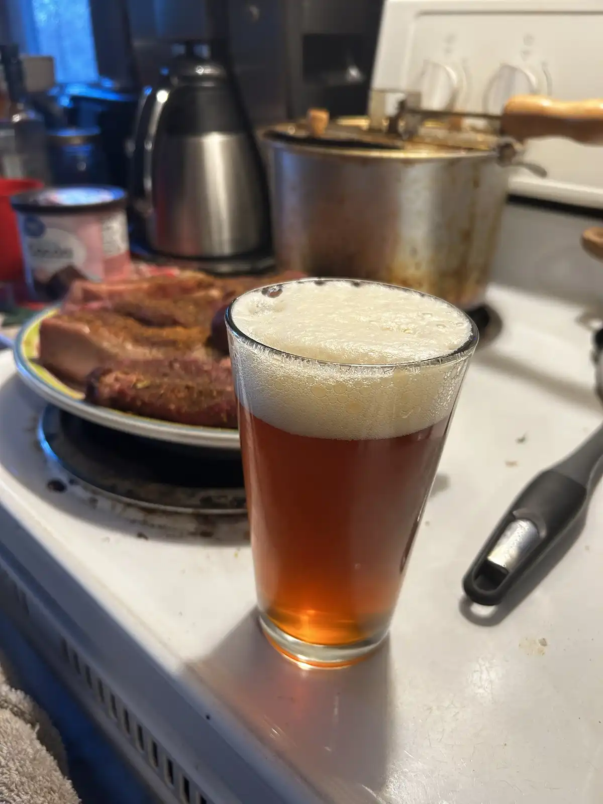 A glass of amber beer with a thick white head sits on a white stovetop next to a plate of seasoned raw meat, with a kettle…