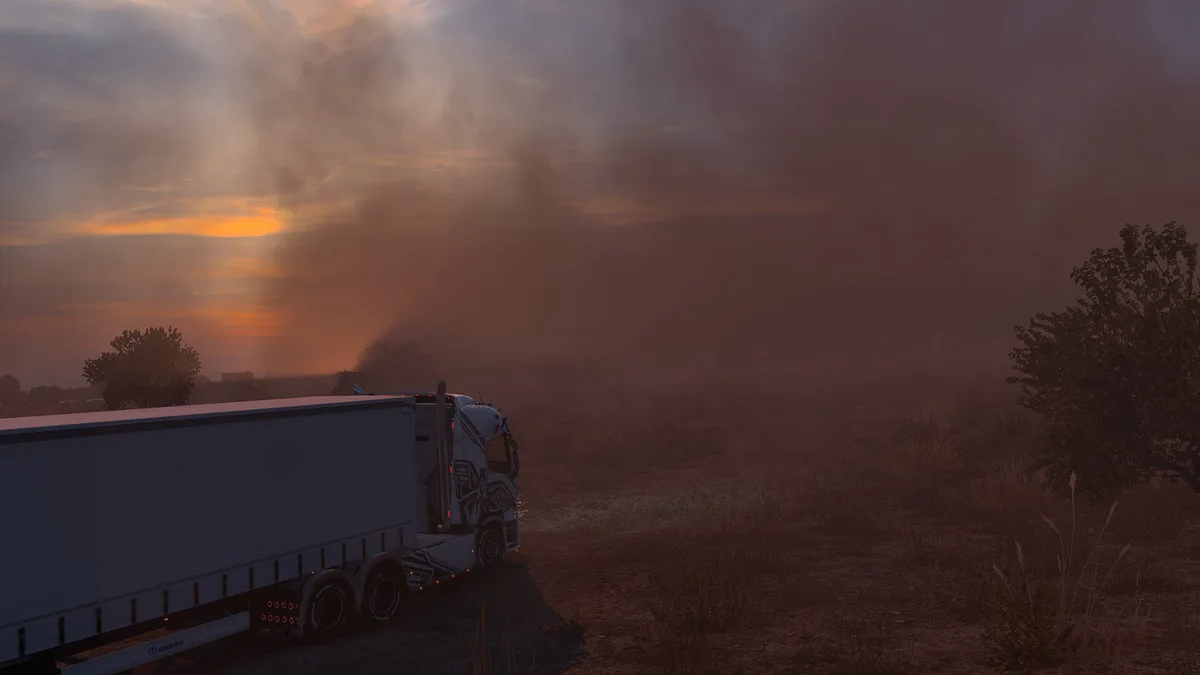 A white semi-truck with a Krone trailer is parked on a dirt road in a field during a dramatic sunset, with a massive…