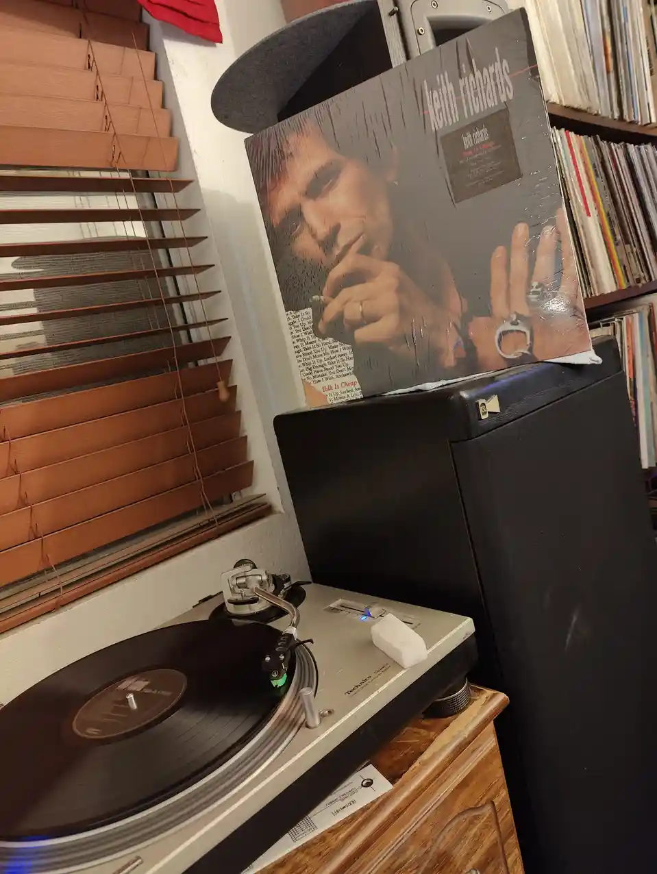 A vinyl record album cover of Keith Richards titled 'Talk Is Cheap' is propped up on top of a black speaker, with a…