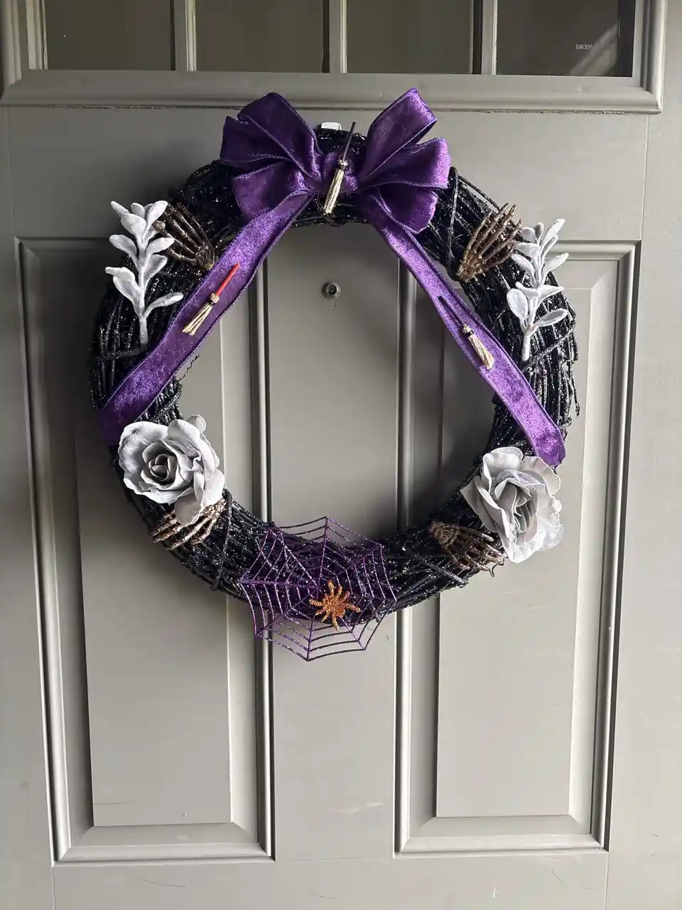 A Halloween wreath hangs on a grey door, featuring a large purple velvet bow, black glittery base, white roses, skeleton…