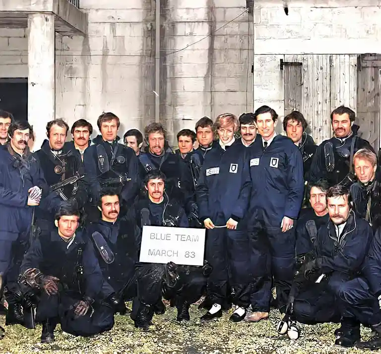 A group photo of men in dark blue jumpsuits posing in front of a concrete structure, one holding a sign that reads 'BLUE…