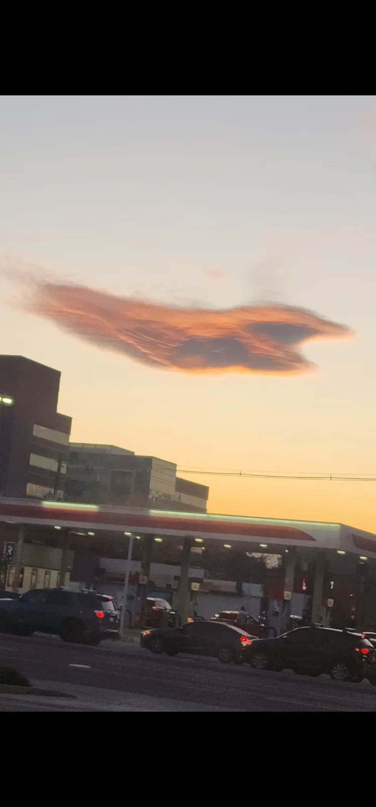 A photo of a gas station at dusk with cars parked and driving by, featuring a long, orange-tinged cloud in the sky that…