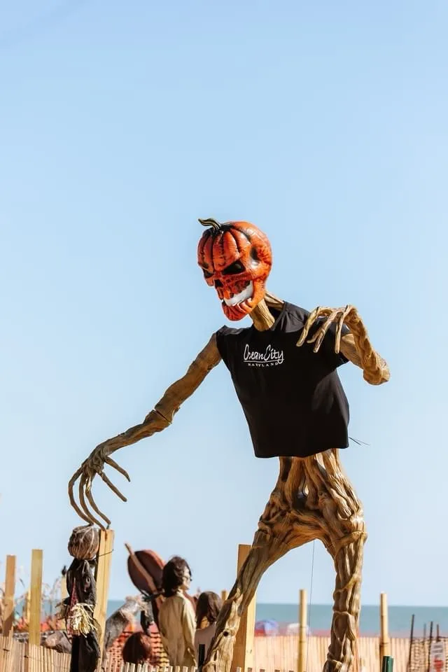 A tall, skeletal scarecrow figure with a carved pumpkin head and twig-like limbs wears a black t-shirt with 'Cream City'…