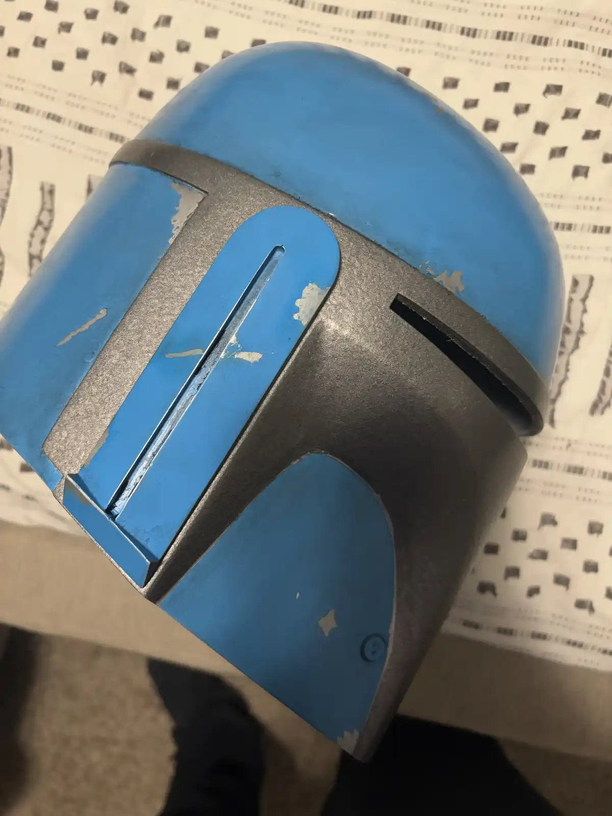A close-up photo shows a blue and grey Mandalorian-style helmet with visible paint chipping and wear, resting on a patterned…