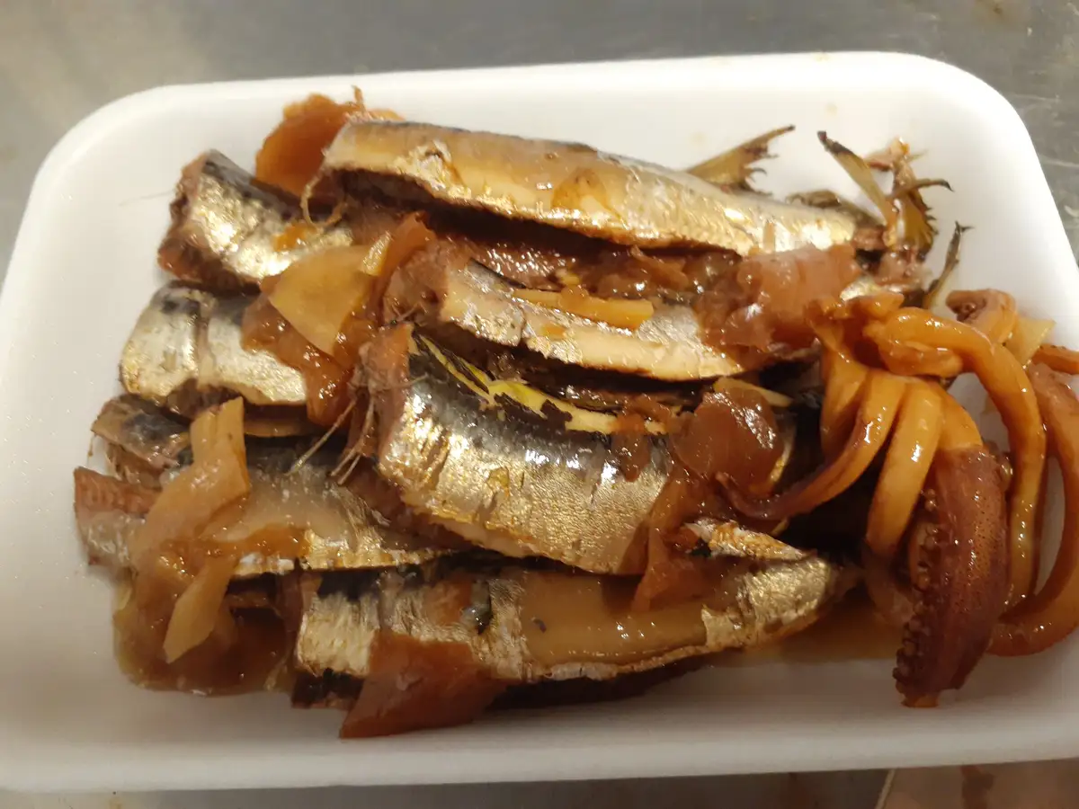 A white styrofoam container holds a pile of cooked fish fillets with shiny silver skin, mixed with caramelized onions and a…