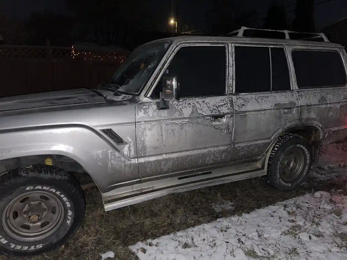 A silver, older model SUV is parked outdoors at night, its entire body covered in a thick layer of snow and ice, with snow…