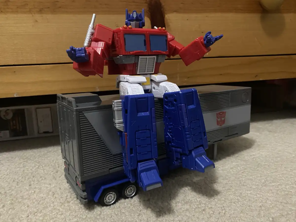 A red, blue, and white Optimus Prime toy robot stands in a heroic pose atop a grey and blue semi-truck toy, with one hand…
