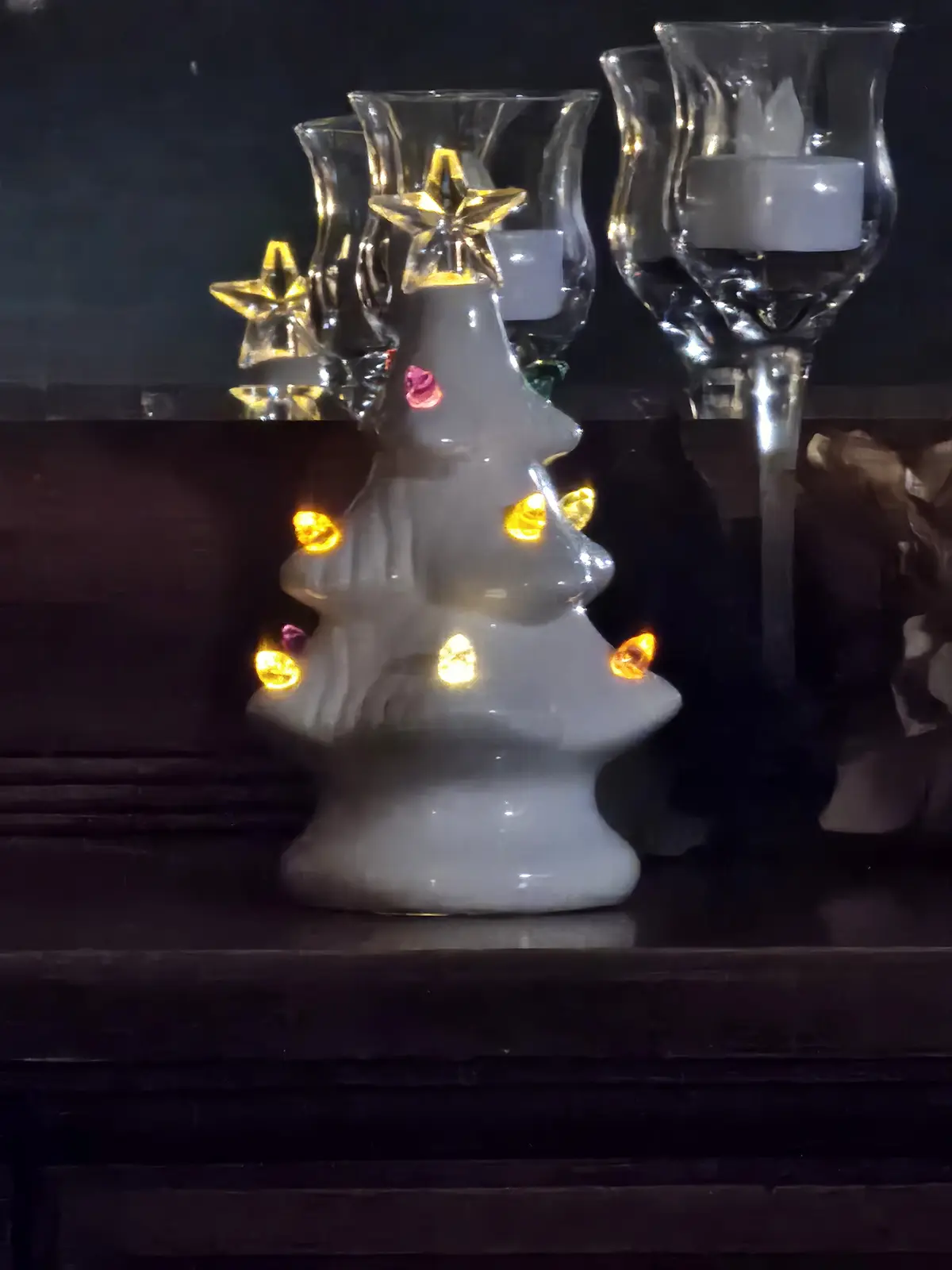 A white ceramic Christmas tree ornament with glowing yellow lights and a pink flower decoration sits on a dark wooden…