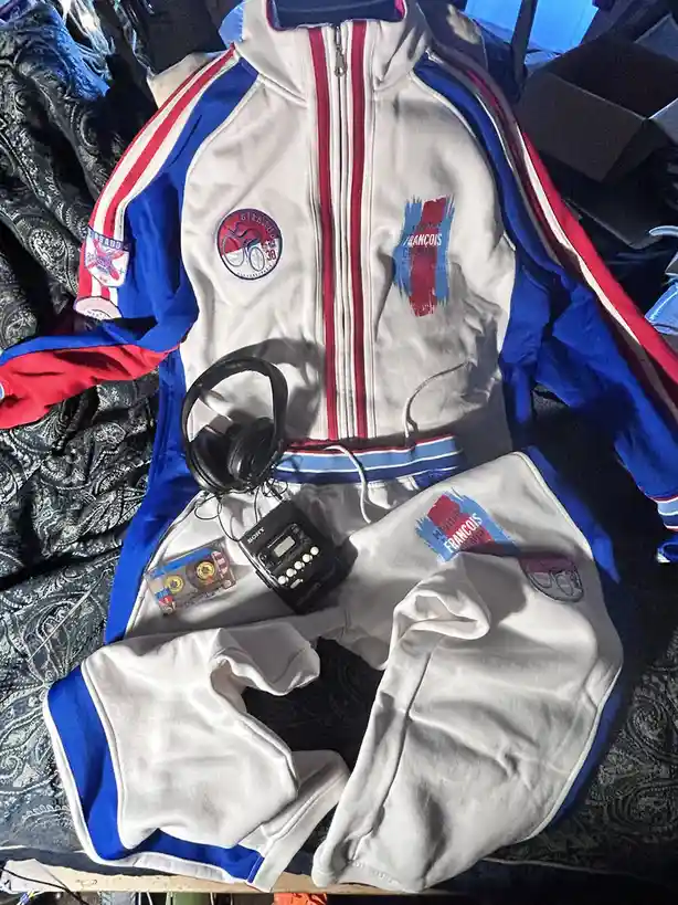 A vintage white, blue, and red track jacket and matching sweatpants are laid out on a patterned fabric surface. Resting on…