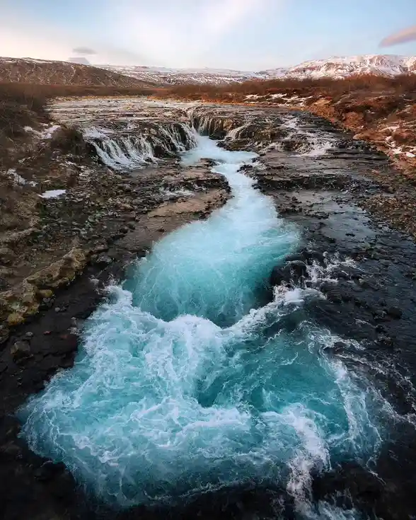 A scenic photograph of a rushing, turquoise river flowing over dark, rocky terrain with snow-dusted hills in the background…