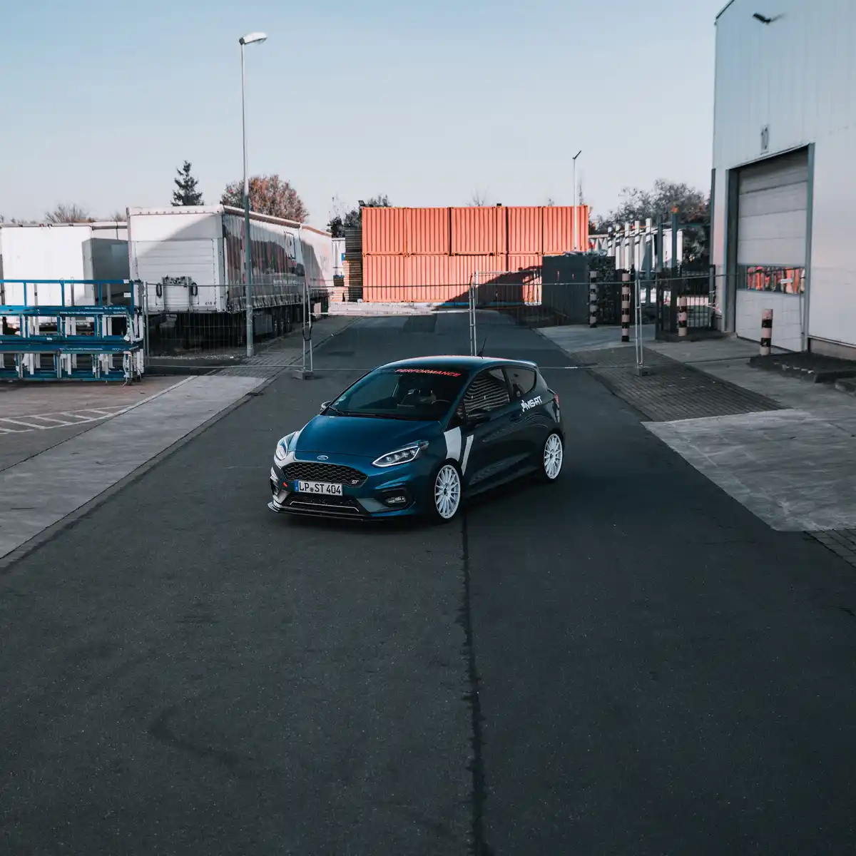 A dark blue Ford Fiesta ST is parked in an industrial lot with shipping containers and warehouses in the background. The car…