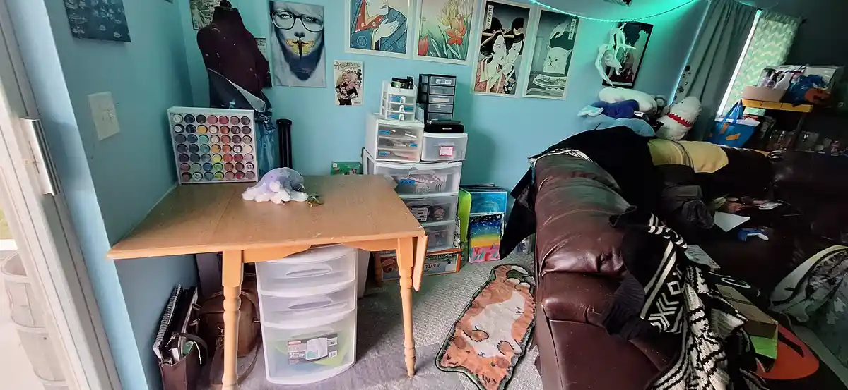 A room with light blue walls, a wooden table, and a brown leather couch covered in blankets and plushies. The table holds a…