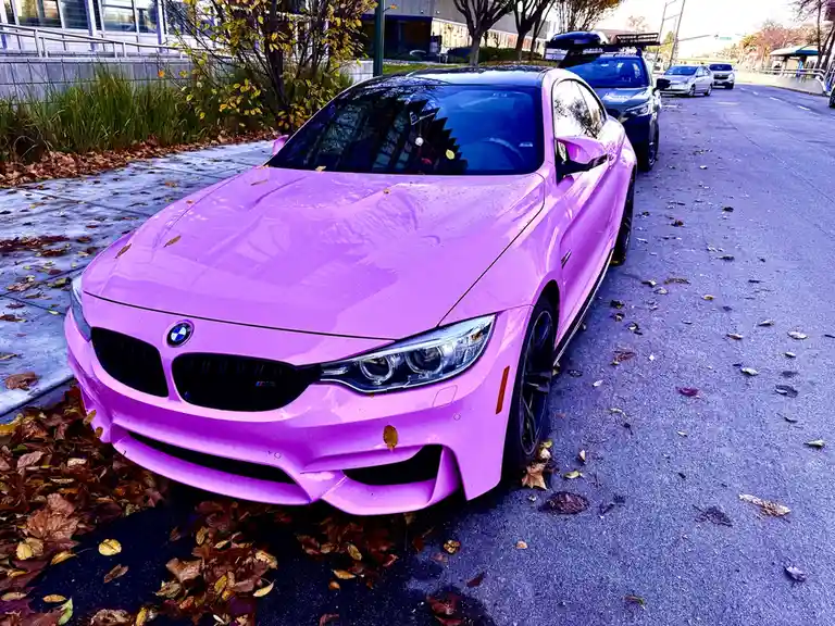 A glossy, bright purple BMW M4 coupe is parked on a street covered with fallen autumn leaves. The car's vibrant color…