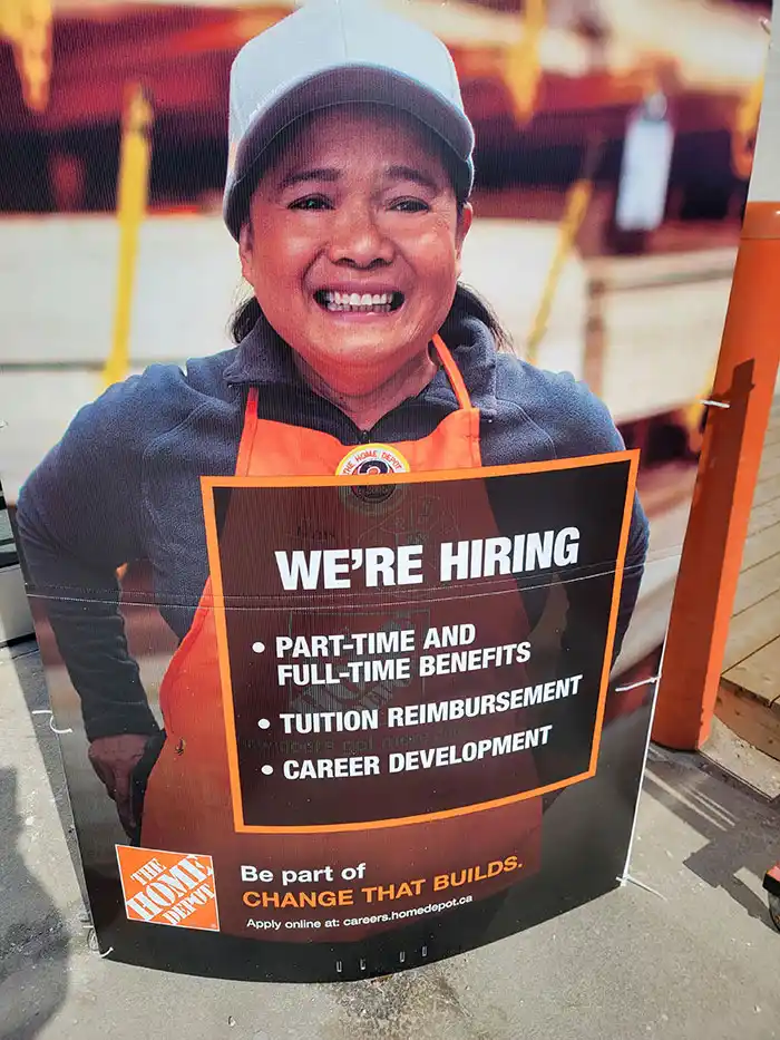 A large, upright Home Depot hiring sign features a smiling woman wearing a white cap and orange apron. The sign lists…