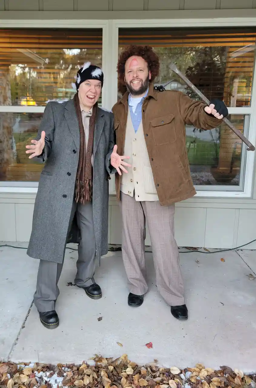 Two people dressed as characters from the TV show 'The Office' stand outside a house. The woman on the left wears a grey…