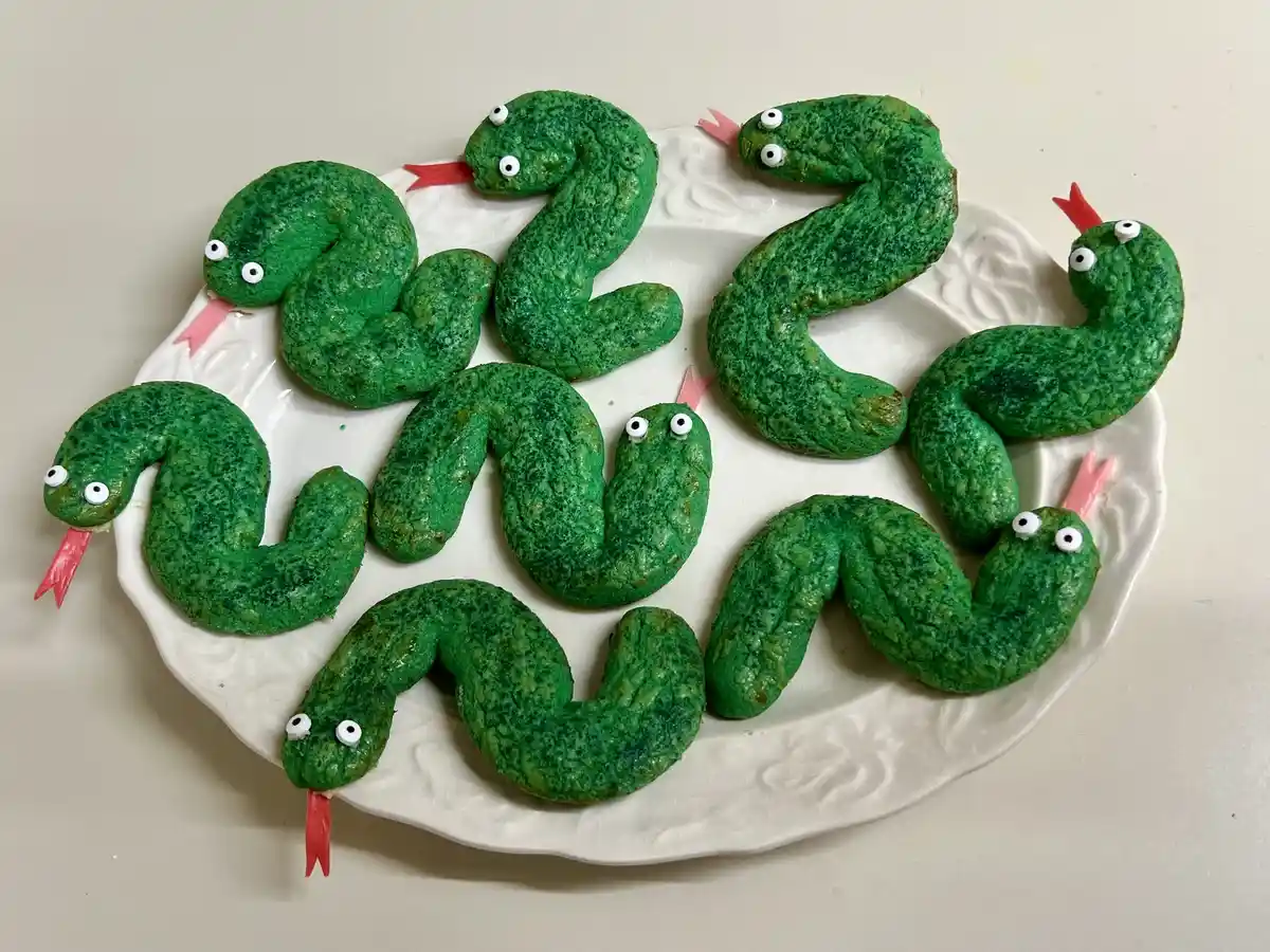 A white plate holds seven green, snake-shaped cookies arranged to spell out the year 2025. Each cookie has googly eyes and a…