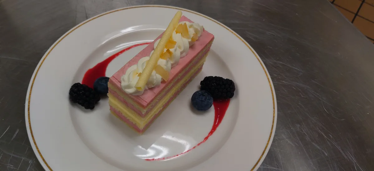 A professionally plated slice of layered cake with pink and yellow layers, topped with whipped cream, a yellow wafer stick…