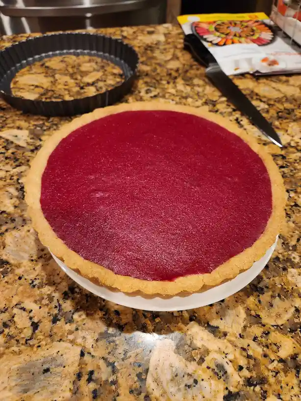 A close-up photo shows a pie with a bright red, smooth filling inside a golden-brown crust, sitting on a white plate atop a…