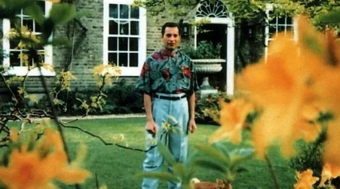A man stands in a garden in front of a brick house with white-trimmed windows, wearing a colorful floral shirt and light…