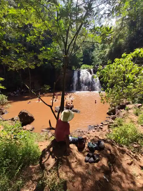 A person in a red shirt and straw hat sits on a rock overlooking a muddy waterfall pool filled with swimmers. Their shoes…