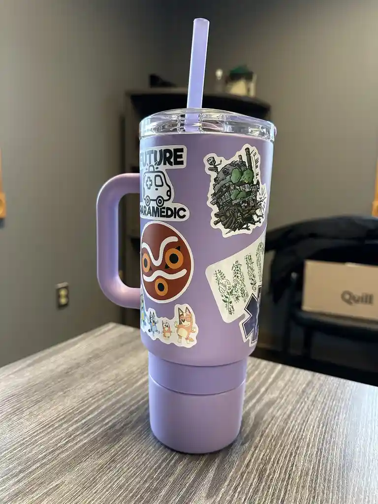 A light purple insulated tumbler with a handle and a clear lid with a purple straw sits on a wooden table. The tumbler is…