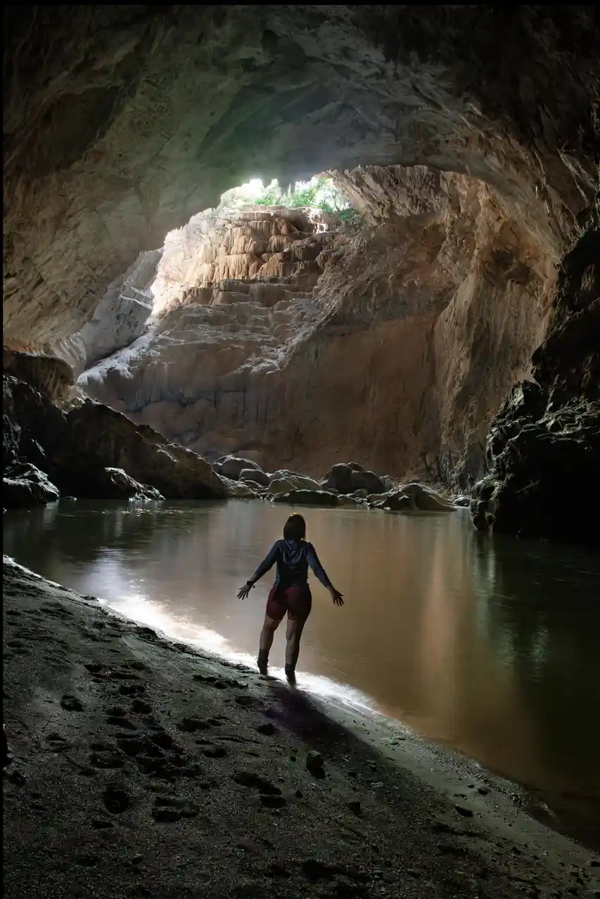 A person stands at the edge of a calm body of water inside a large, dimly lit cave, facing a bright opening where sunlight…