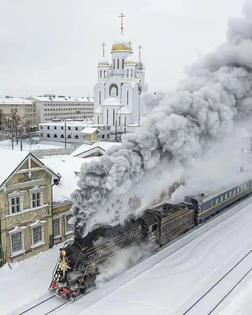 A steam train emits thick black smoke as it travels through a snowy town, passing a prominent white Orthodox church with…