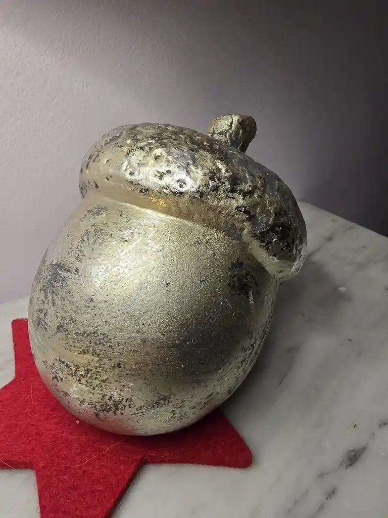 A gold-painted, slightly worn acorn-shaped decorative object sits on a red felt star atop a marble surface. The overlaid…