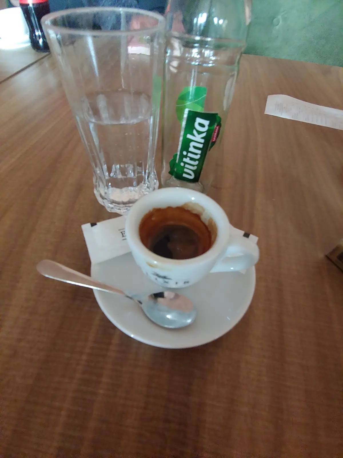 A nearly empty espresso cup sits on a saucer with a spoon, next to a glass of water and an empty 'Vitinka' bottle on a…