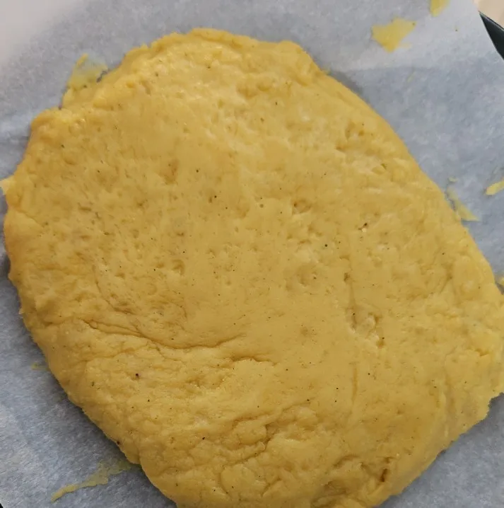 A close-up photo shows a flattened, yellowish, dough-like substance on parchment paper, with small dark specks scattered…