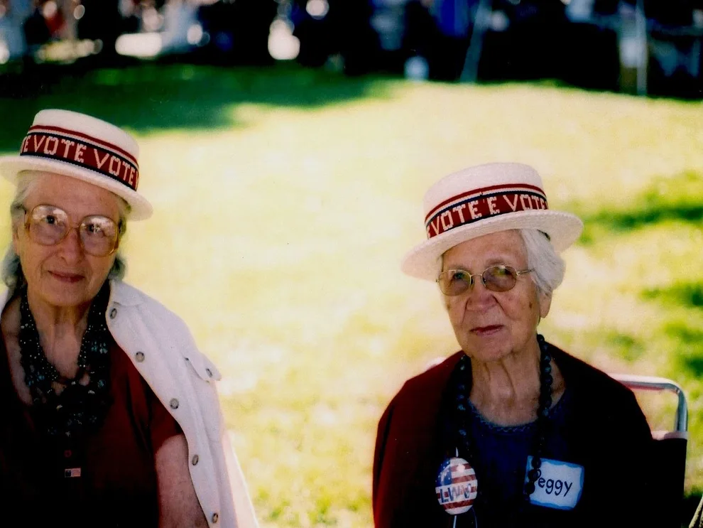 Two elderly women wearing matching white hats with red "VOTE E VOTE" bands sit outdoors on a grassy field. The woman on the…