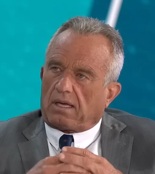 a close-up of a middle-aged man with gray hair, wearing a gray suit and blue tie, looking concerned while clasping his…