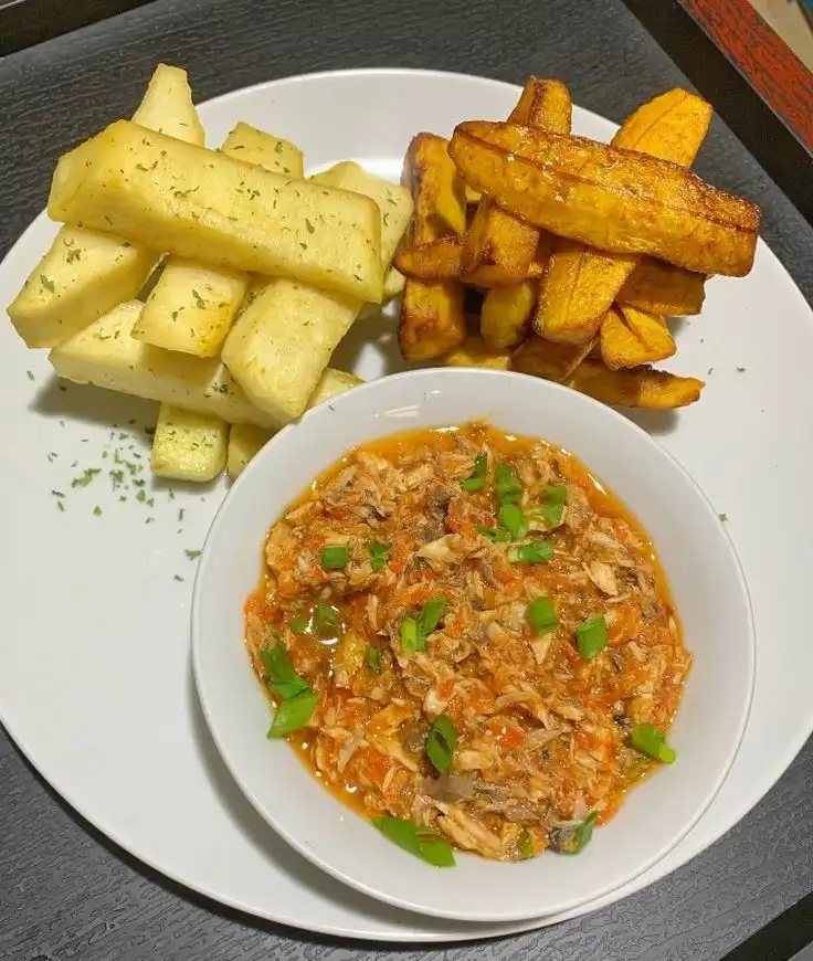 A white plate holds a bowl of shredded meat in a tomato-based sauce garnished with green onions, alongside two types of…
