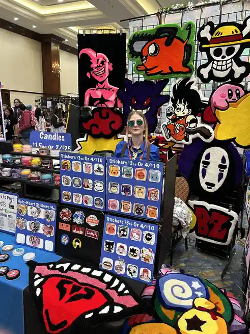 A woman wearing sunglasses stands behind a vendor table at a convention, surrounded by large, colorful, fluffy rug-like…