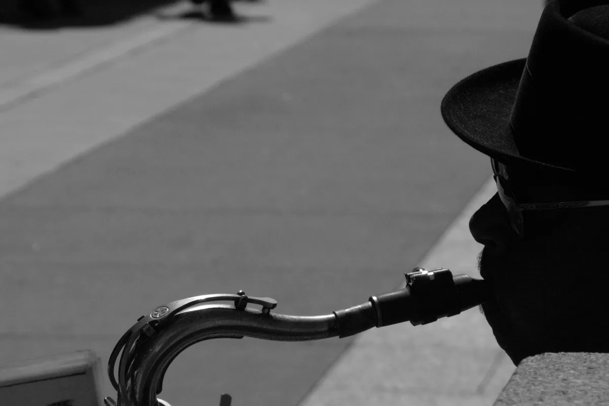 A black-and-white photo shows a person in profile, wearing a fedora and sunglasses, playing a saxophone on a sidewalk. The…