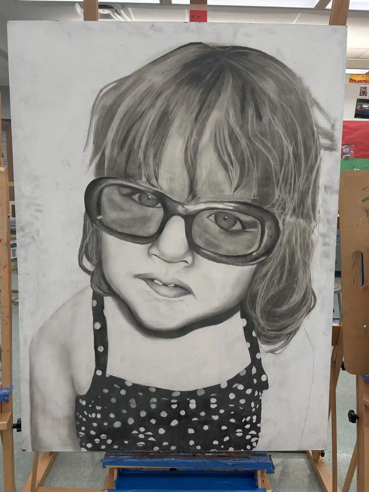 A large, detailed pencil drawing on a canvas shows a young child with thick-rimmed glasses and a polka-dot top, mounted on…