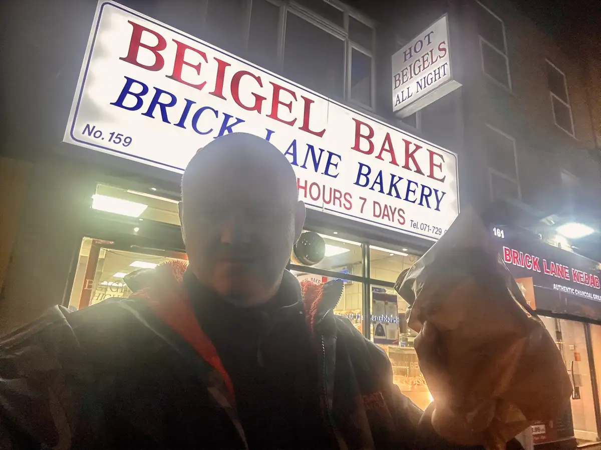 A man takes a selfie at night outside 'Beigel Bake Brick Lane Bakery', holding a beigel in a paper bag. The bakery sign…