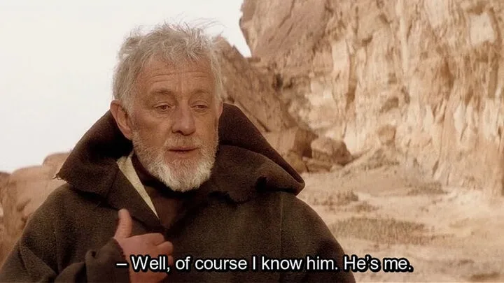 An older man with a white beard and gray hair, wearing a brown hooded robe, stands in a desert landscape with rocky cliffs…