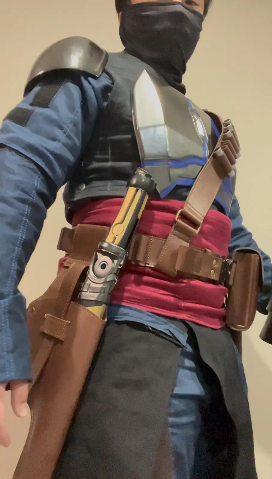 A person in a Star Wars cosplay costume, wearing a black face mask, blue shirt, and grey armor with a red sash, has a…