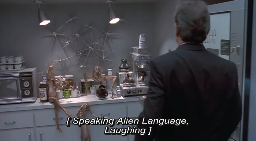 A man in a suit stands with his back to the camera in a kitchen, looking at three alien creatures on the counter. The aliens…