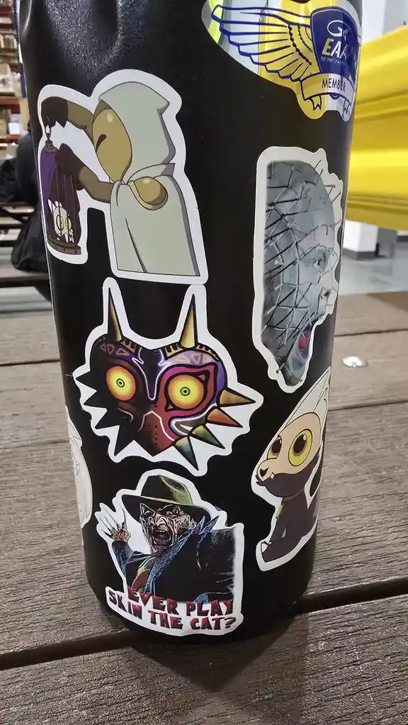 A black tumbler covered in various stickers, including a hooded figure holding a cage, a menacing mask, a cracked skull, a…