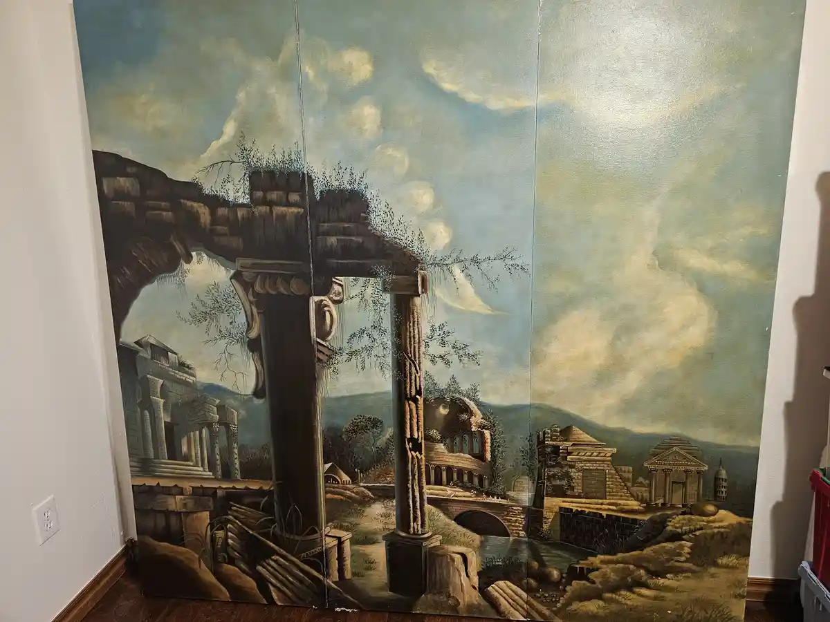 A large, three-panel painting of classical ruins and ancient architecture is leaning against a wall in a room, with visible…