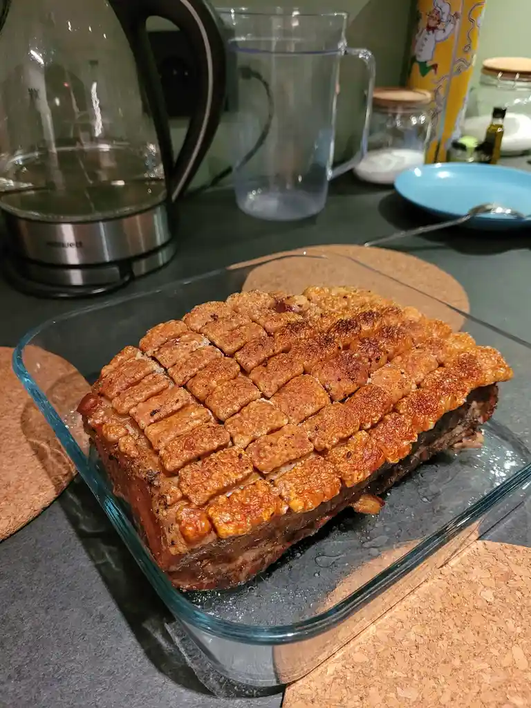 A rectangular piece of roasted meat with a golden-brown, crispy, grid-patterned crust sits in a clear glass baking dish on a…